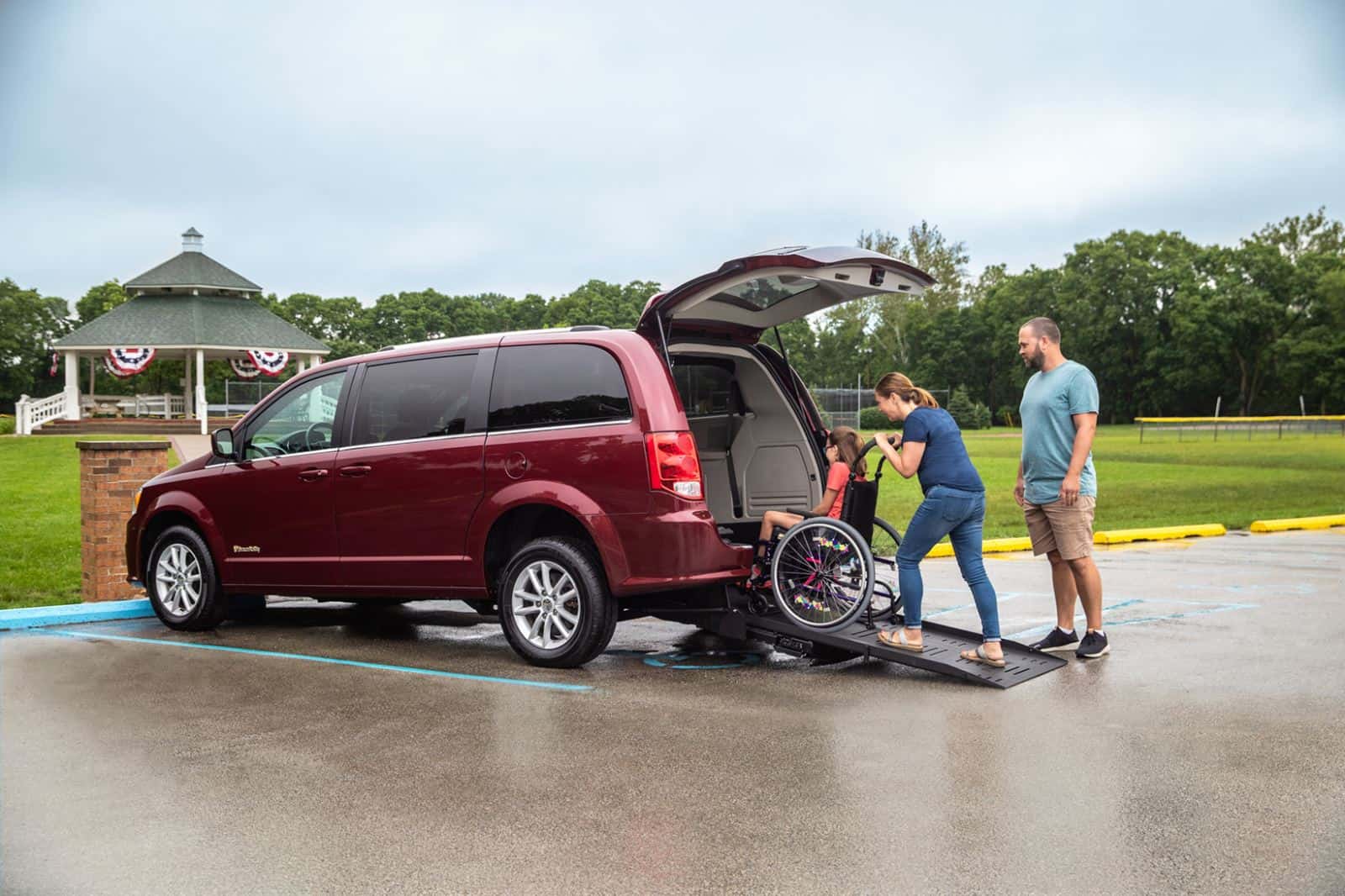 a man watches as a woman rolls a young girl up a ramp into a red van